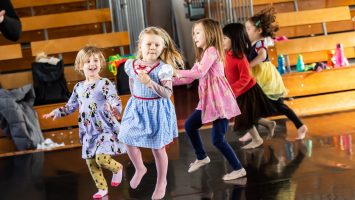 A group of young children wearing colourful dresses skip in a group during a dance class.