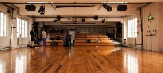 Four artists chat at the back of a large dance studio with shiny wooden floors and a bank of raised seating
