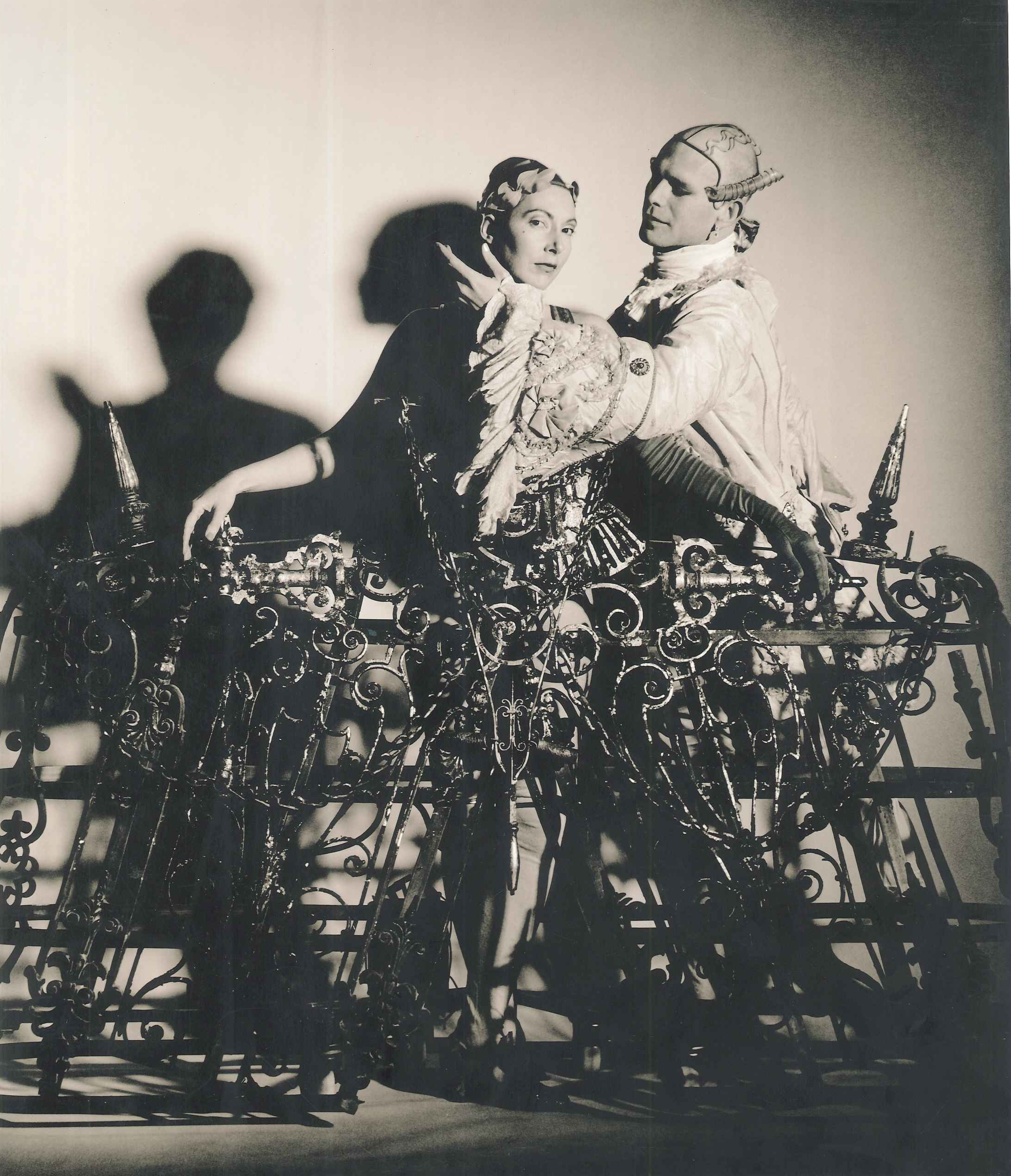 A sepia image of two performers dressed in ornate ruffled costumes stand delicately inside an elaborate waist-height cage