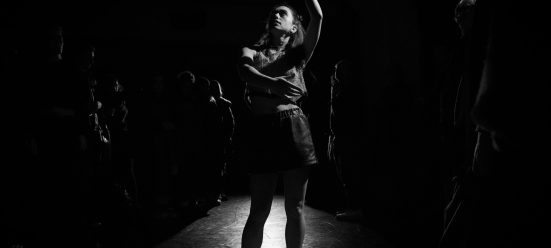 A light-skinned performer stands with curved arms raised upwards, on a shiny dance floor. They are surrounded by a standing audience in almost total darkness