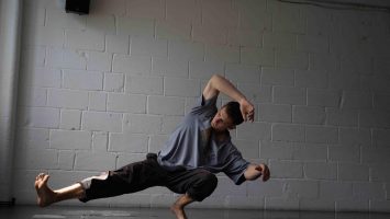 A dancer stretches his right leg out while crounching. He has a blue tshirt and black trousers. There is a white brick wall behind and a black dance floor.