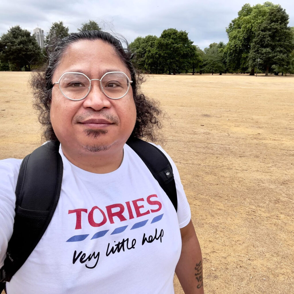 An image of Michael in front of a yellow field and green trees. Michael is wearing glasses, and has curly black hair. He is wearing a white t-shirt that says ‘Tories - very little help'